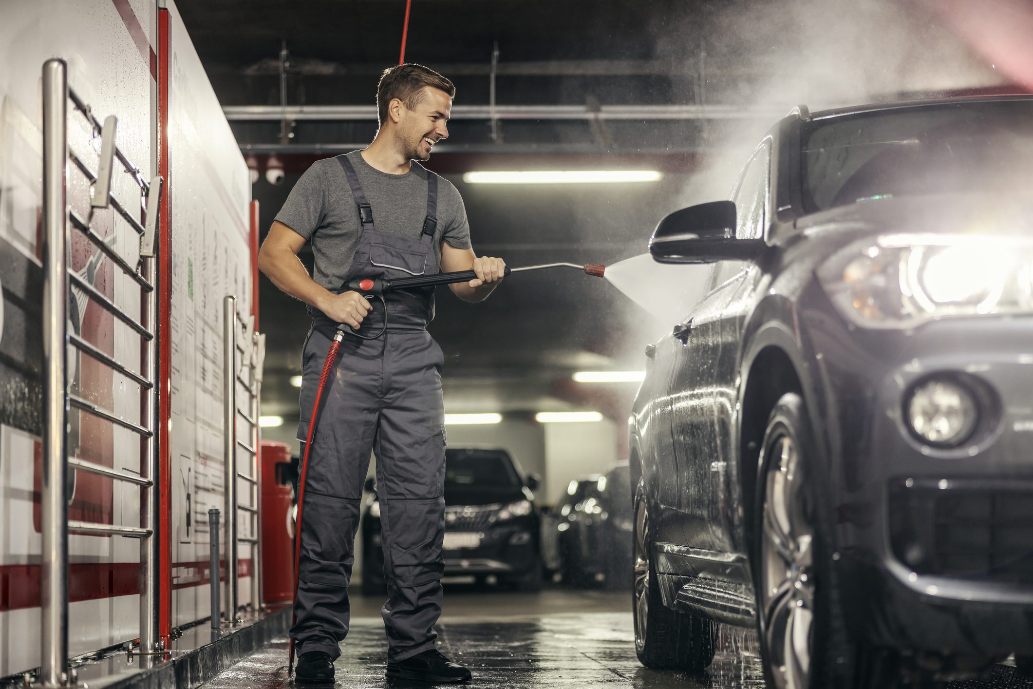A cheerful car wash worker is washing a car with water jet gun at station.