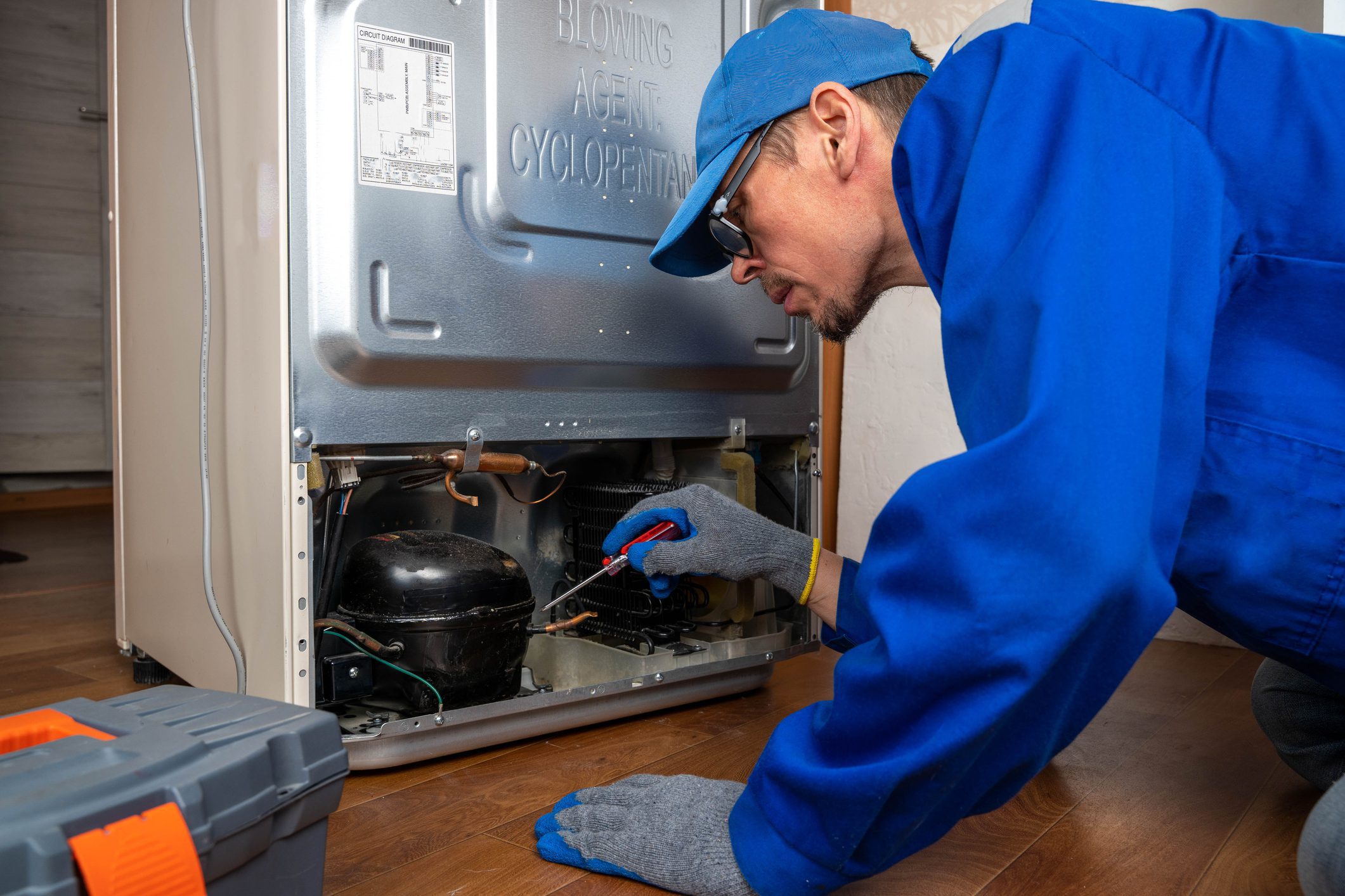 Repair work takes place on a refrigerator in a kitchen setting.