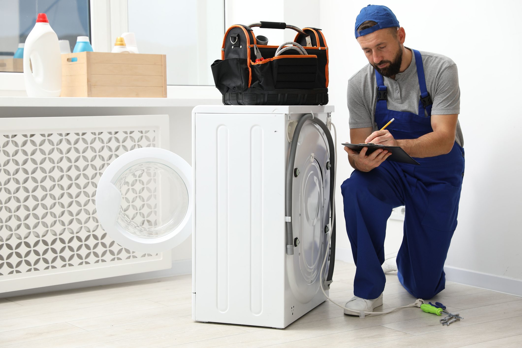 Professional repairman with clipboard and bag of tools near broken washing machine indoors